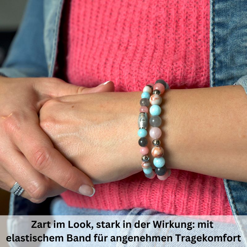 Eine Person trägt zwei farbenfrohe Armbänder, darunter das Armband Pellworm mit Pink Jade, Quarz-Kristall, Rotem Netz Jaspis und Edelstahl-Beads, über einem rosa Strickpullover. Der deutsche Text beschreibt das filigrane Design und das bequeme Elastikband.