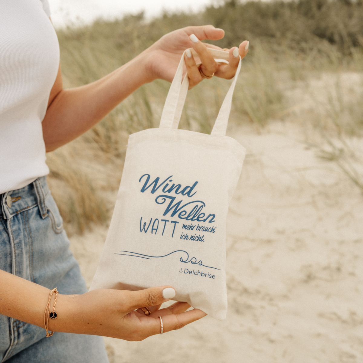 Frau hält die Deichbrise Tragetasche mit dem Spruch Wind, Wellen, Watt am Strand