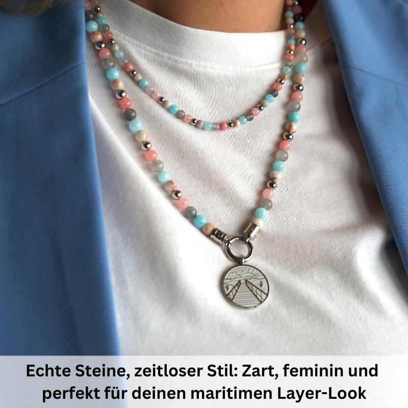 Close-up of a person wearing two beaded necklaces, including the Kette Pellworm (4mm) aus Pink Jade, Quarz-Kristall und Roter Netz Jaspis with pastel stones and round pendant, paired with a white shirt and light blue blazer. Timeless, feminine layered look.