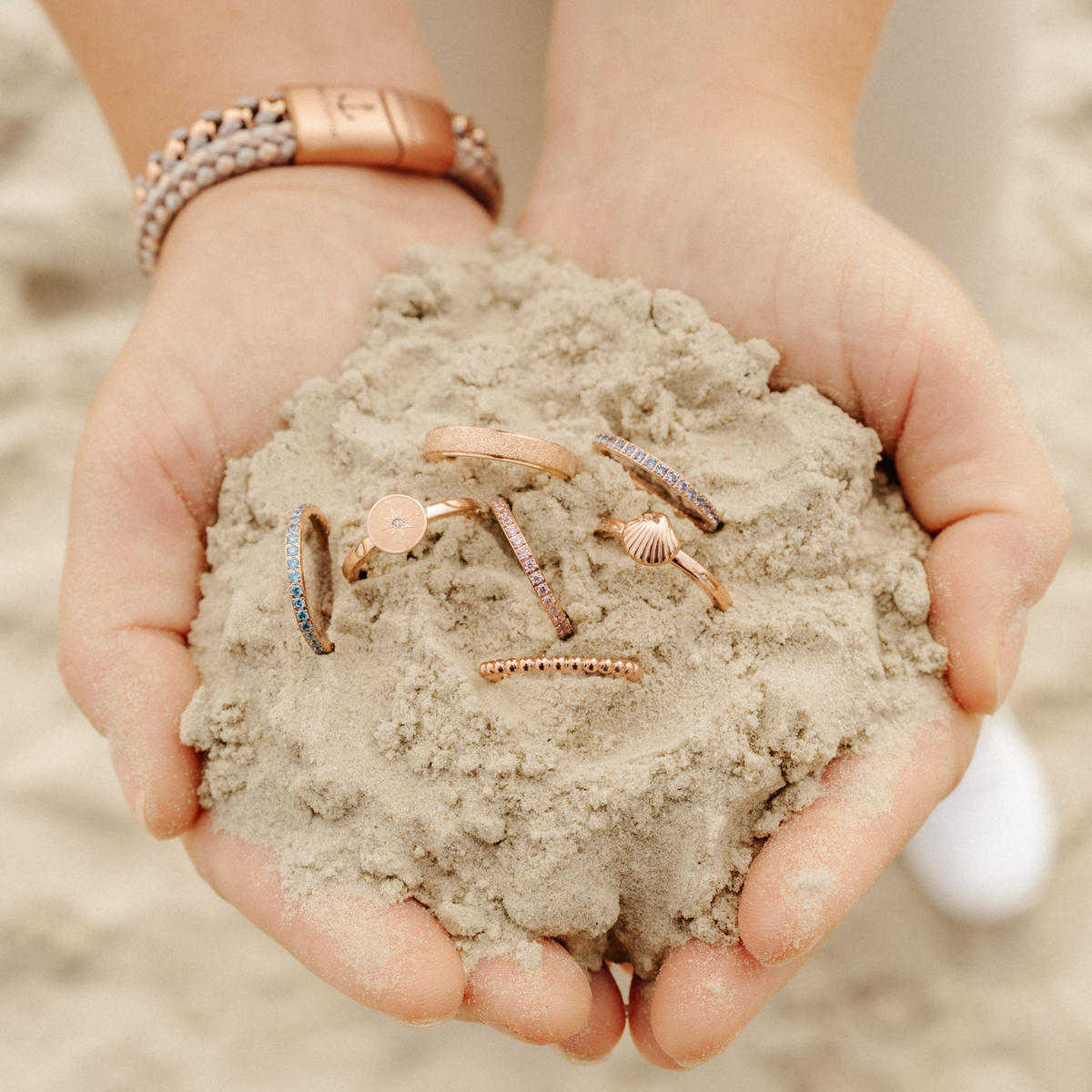 Hände voller Sand mit verschiedenen Stapelringen aus Edelstahl, darunter der Ring Bling Bling