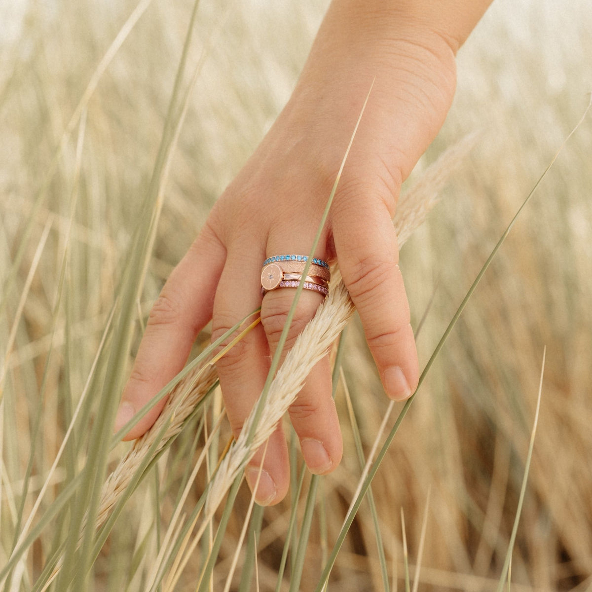 Hand im Dünengras mit gestapelten Ringen, darunter der Ring Bling Bling mit rosa Steinen