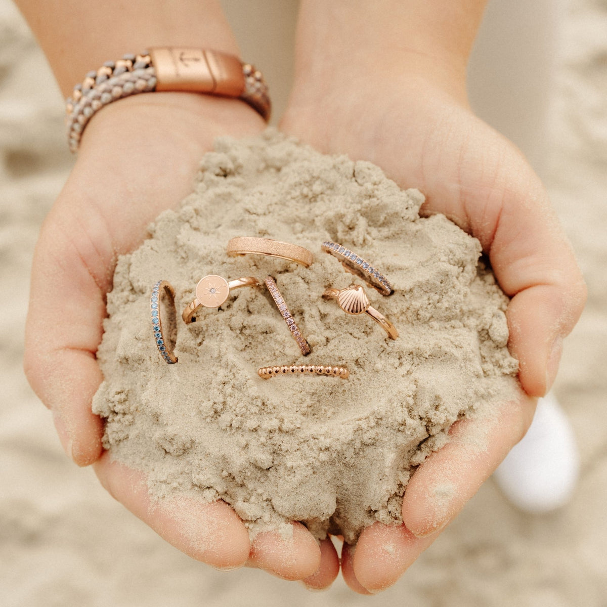 Hände voller Sand mit verschiedenen Stapelringen aus Edelstahl in Roségold