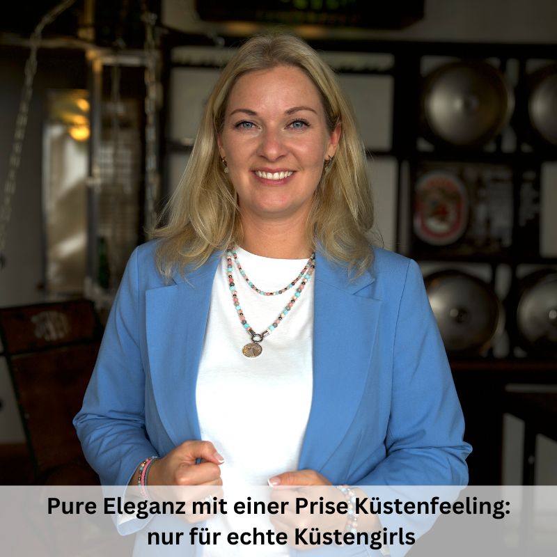 A smiling blonde woman indoors wears a light blue blazer, white top, and the Kette Pellworm (4mm) aus Pink Jade, Quarz-Kristall und Roter Netz Jaspis. Text below: Pure Eleganz mit einer Prise Küstenfeeling—nur für echte Küstengirls.